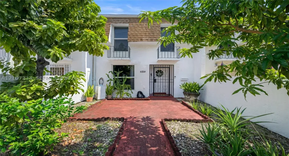Exterior facade featuring white stucco finish, second-story decorative iron balcony railings, a metal security door, and a painted masonry walkway