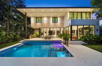 Modern two-story exterior featuring floor-to-ceiling glass sliding doors, a flat roofline, an integrated swimming pool with a stone patio surround, and a second-story balcony with a metal railing
