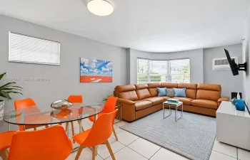 Open-concept room featuring light-colored tile flooring, a bay window with horizontal blinds, neutral wall paint, a ceiling-mounted flush light fixture, and a wall-mounted climate control unit