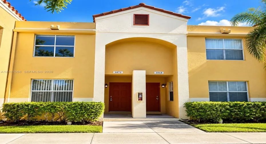 Stucco exterior featuring a dual-entry arched entryway, terra cotta-toned roof tiles, manicured shrubbery, and concrete walkways
