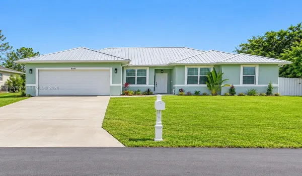 Single-story residence featuring a light-colored metal roof, pale green stucco siding, a two-car garage, and a concrete driveway