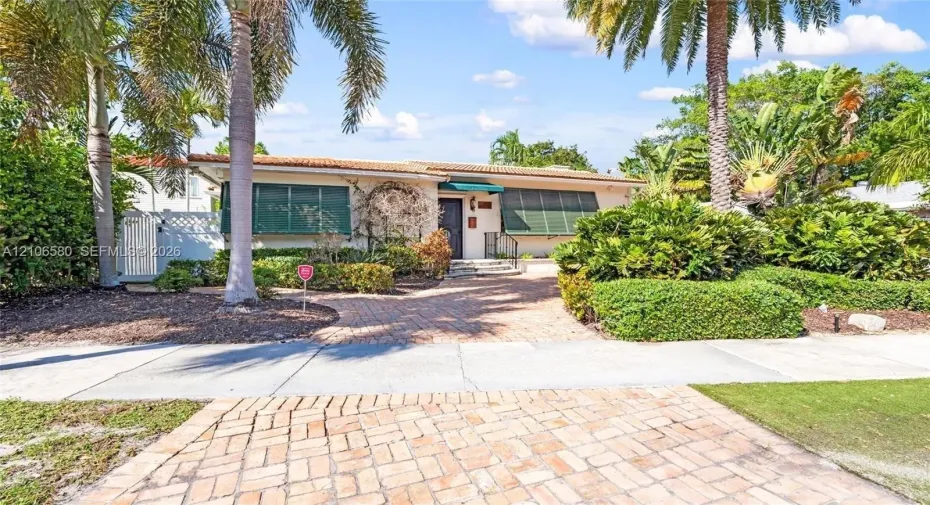 Single-story residence featuring a brick-paved driveway, clay tile roof, metal window awnings, and mature landscaping