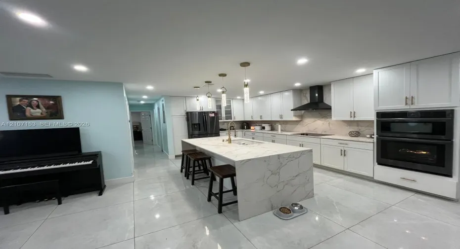 Kitchen featuring white cabinetry, waterfall-edge island with stone-finish countertop, stainless steel double wall oven, patterned stone-finish backsplash, and recessed lighting