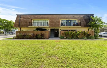 Two-story exterior featuring a shingled mansard roof, stucco siding, dual upper-level balconies with black metal railings, and a fenced ground-level perimeter