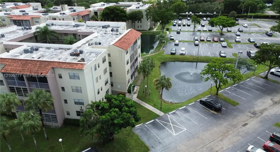 Multi-story residential building featuring a terracotta tile roof, balcony enclosures, landscaped grounds, a central fountain, and dedicated parking areas