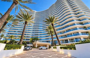 Multi-story architectural exterior featuring a curved facade, expansive balconies with glass railings, patterned stone paver motor court, and manicured landscaping with mature palms