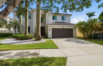 Two-story residence featuring a light-toned exterior, concrete tile roof, multi-car garage, and landscaped front grounds with mature palm trees
