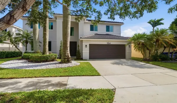 Two-story residence featuring a light-toned exterior, concrete tile roof, multi-car garage, and landscaped front grounds with mature palm trees