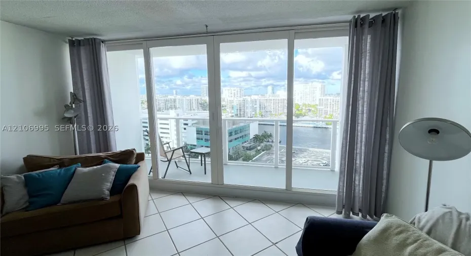 Floor-to-ceiling glass sliding doors, private balcony with glass railing, ceramic tile flooring, and urban water scenery