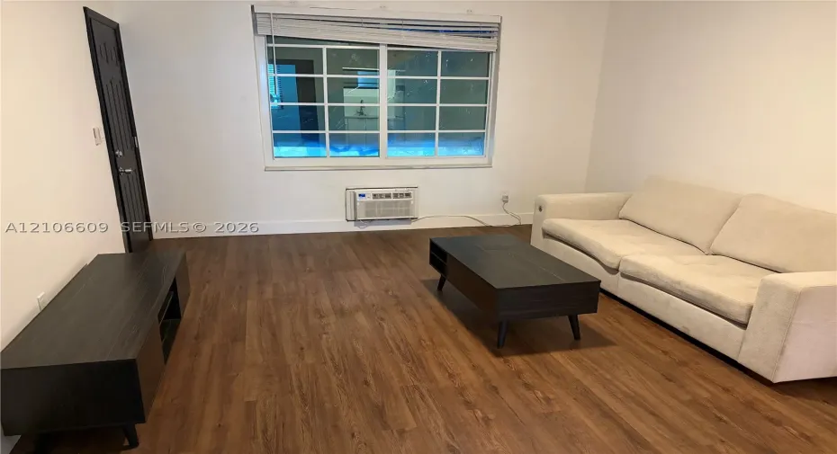 Wood-finish flooring, multi-pane window with top-down blinds, and wall-mounted climate control unit