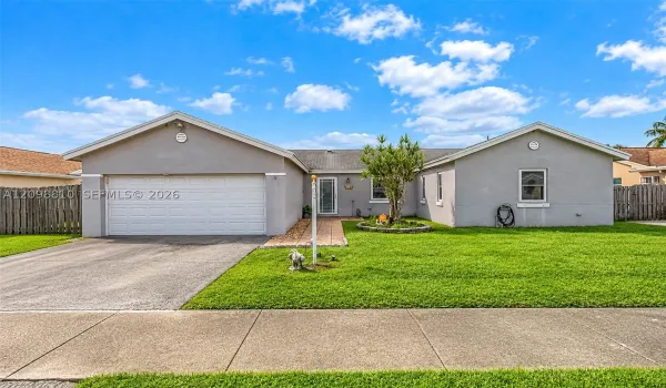 Single-story residence featuring a gray stucco exterior, a two-car garage, a paved walkway, and a landscaped front yard with grass lawn and wood-style fencing