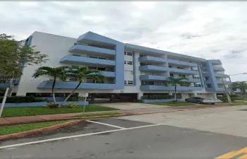 Multi-story residential building featuring blue-accented exterior, private balconies, covered ground-level parking, and landscaped grounds