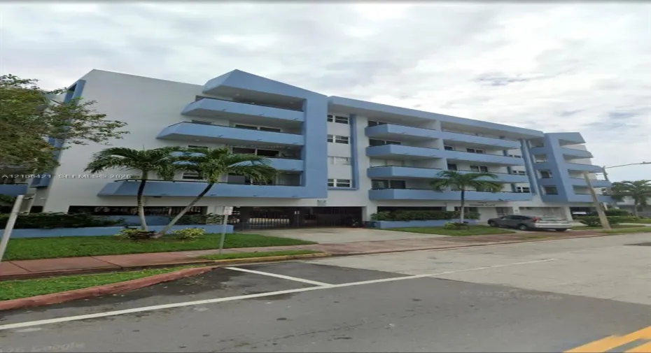 Multi-story residential building featuring blue-accented exterior, private balconies, covered ground-level parking, and landscaped grounds