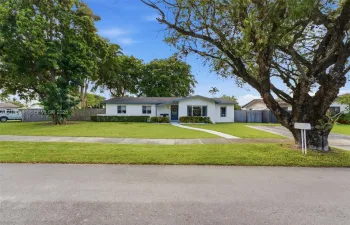 Single-story residence featuring light-colored exterior siding, a wide front lawn, mature shade trees, a concrete walkway, and private perimeter fencing