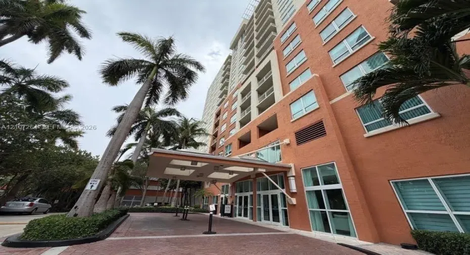 High-rise exterior featuring a structured porte-cochère, textured masonry facade, multi-story glass fenestration, and landscaped palm trees along a paver stone arrival court
