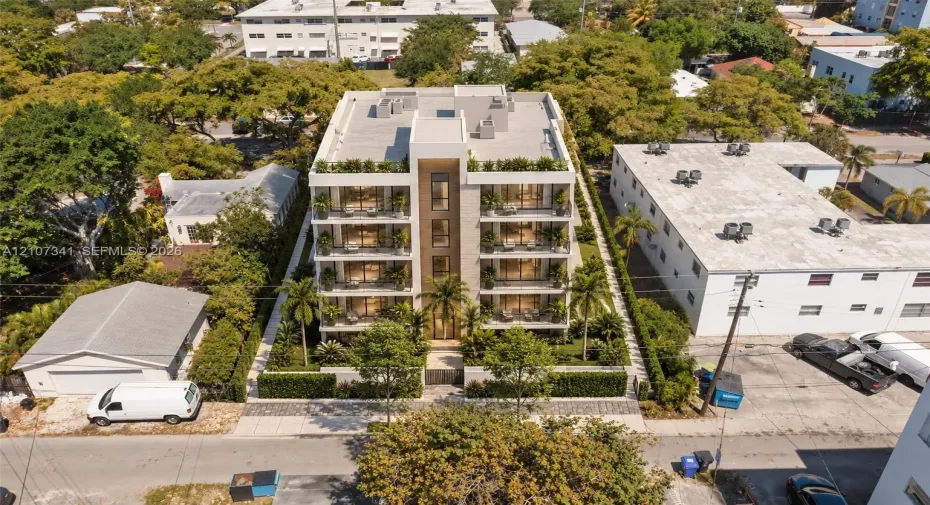 Multi-story residential building featuring a stone-clad facade, private balconies on each level, floor-to-ceiling windows, and landscaped perimeter hedging