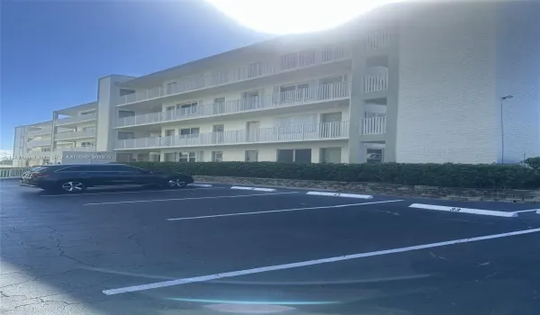 Multi-story building exterior featuring white brick siding, horizontal metal balcony railings, and surface lot parking