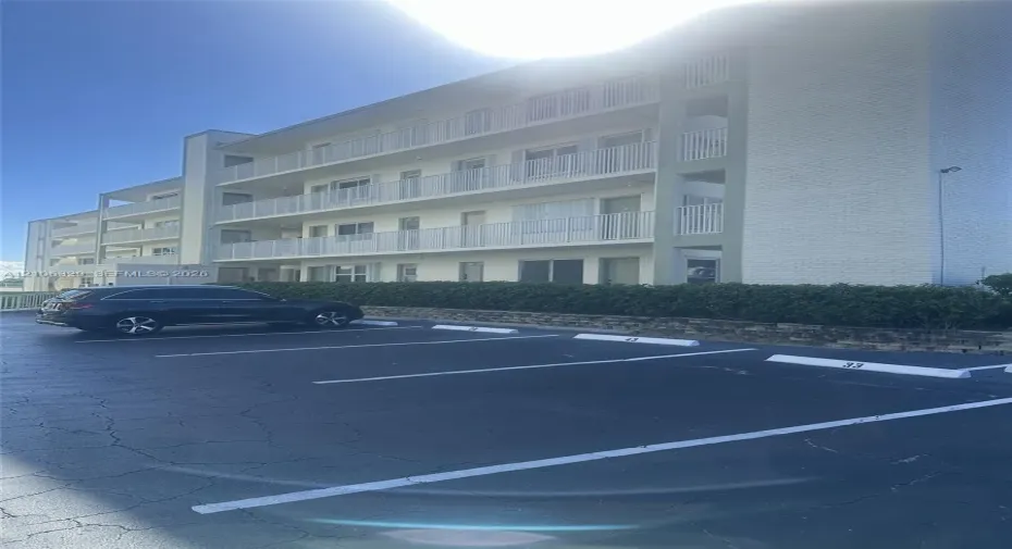 Multi-story building exterior featuring white brick siding, horizontal metal balcony railings, and surface lot parking