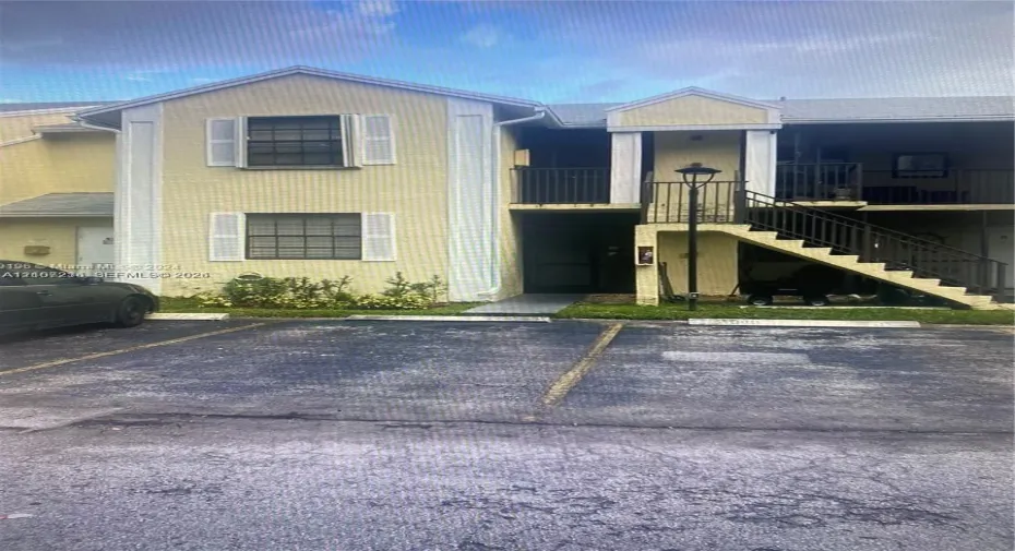 Two-story multi-family exterior featuring light-toned stucco siding, louvered shutters, metal railings, and a dedicated staircase