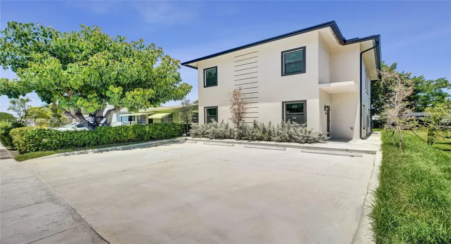 Two-story exterior featuring light-toned stucco siding, dark-trimmed windows, a concrete parking area, and mature landscaping