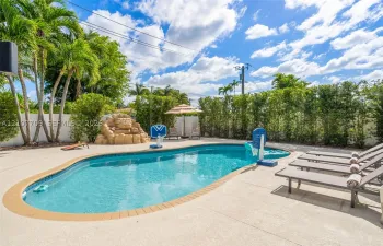 In-ground swimming pool featuring a brick-tiled border, decorative stone fountain, concrete pool deck, and privacy hedging