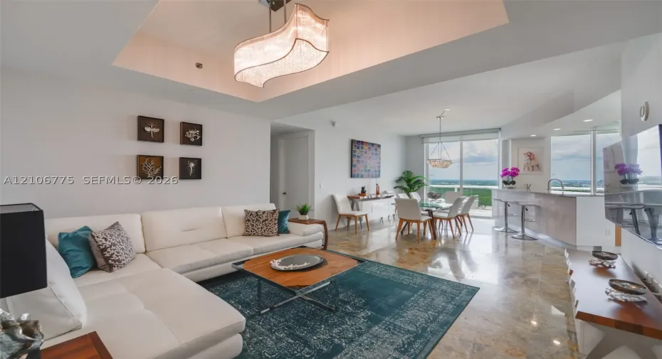 Open-concept living space featuring high tray ceilings with an oversized modern chandelier, polished stone flooring, a built-in kitchen peninsula with stone countertops, and floor-to-ceiling windows