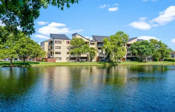 Multi-story residential building featuring a light-toned stucco exterior, pitched metal roofing, private balconies, and mature landscaping along a waterfront setting
