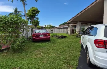 Exterior of the property featuring a covered porch, white vertical-slat fencing, mature palm trees, and manicured green space