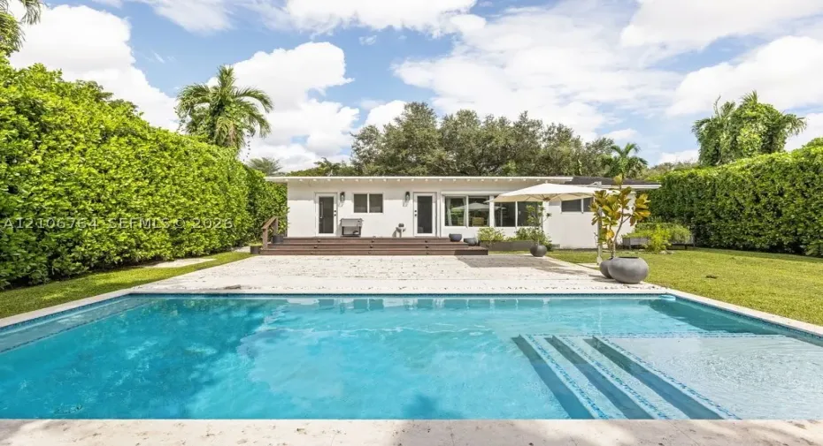 Outdoor area featuring a swimming pool with integrated steps, stone-finish pool deck, elevated wood-finish deck, and surrounding manicured privacy hedges