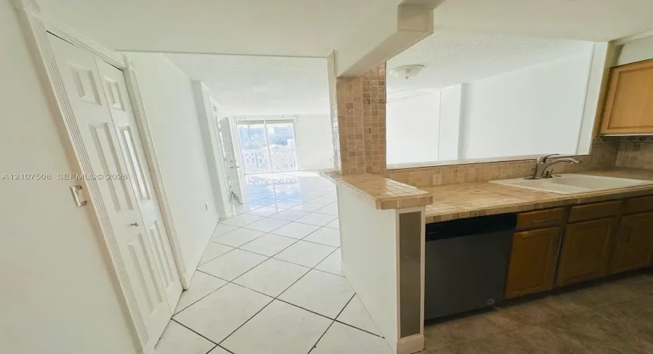 Kitchen featuring tile countertops, mosaic tile backsplash, wood-finish cabinetry, a built-in dishwasher, and white ceramic tile flooring