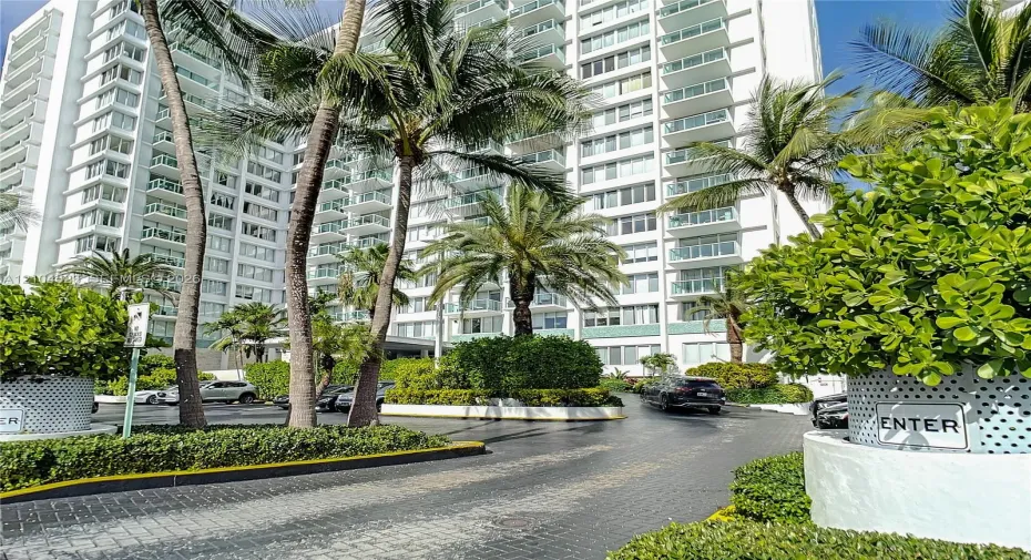 High-rise residential exterior featuring white concrete facade, stacked glass-railed balconies, paved circular motor court, and established tropical landscaping