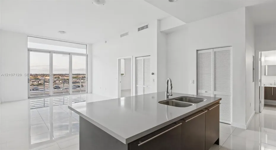 Open-concept kitchen featuring an island with a stone-composite countertop, dual-basin stainless steel sink, and high-arc faucet, complemented by floor-to-ceiling windows and polished tile flooring