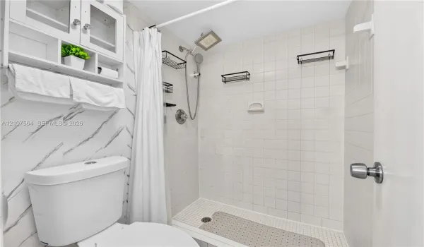 Bathroom featuring a shower stall with white tile surround, a built-in overhead showerhead with an adjustable handheld attachment, wall-mounted shelving, and white cabinetry