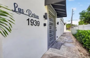 Exterior facade featuring smooth stucco finish, custom metal address signage, wall-mounted cylindrical sconces, dark metal awning, and louvered entry door