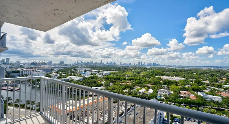 Private balcony with metal railings overlooking an expansive cityscape, waterway, and dense tree canopy