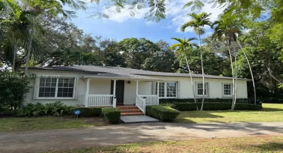 Single-story residence featuring a covered front porch with a white balustrade, brick entry steps, multi-pane windows, and manicured landscaping with mature palm trees