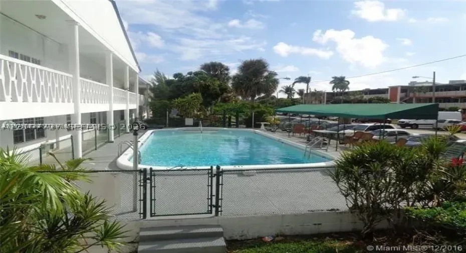 Rectangular swimming pool featuring a perimeter fence, a metal gate, and a covered patio area