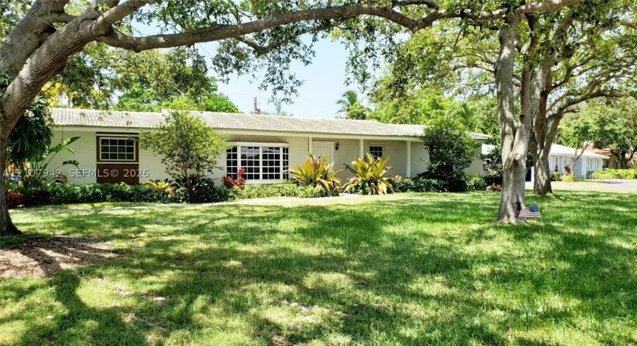Single-story exterior featuring a light-toned stucco façade, tile roof, and bay window, complemented by expansive grounds with mature shade trees