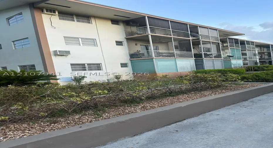 Multi-story building exterior featuring screened balconies with metal railings, horizontal sliding windows, and exterior mounted air conditioning units, complemented by a stone-mulched landscape border with mature shrubbery