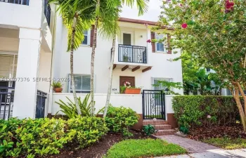 Stucco exterior featuring a second-story balcony with metal railing, a gated brick walkway entrance, and mature palm landscaping