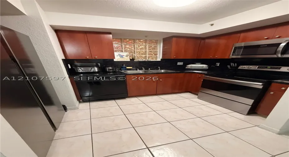 Kitchen featuring wood-finish cabinetry, stainless steel appliances, dark stone-style countertops, ceramic tile flooring, and glass block accent window