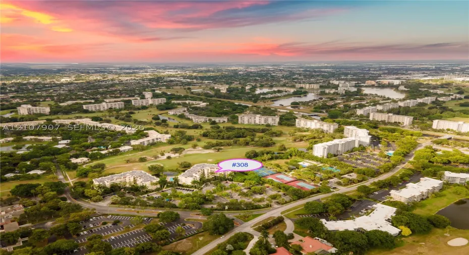 Aerial perspective of a multi-story residential building featuring on-site tennis courts, surface parking, and extensive surrounding green space