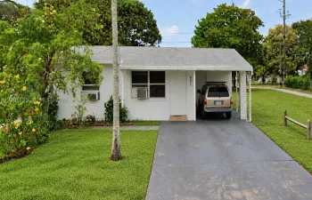 Single-story exterior featuring a light-toned facade, gabled roofline, concrete driveway, and covered carport with stone pillar support