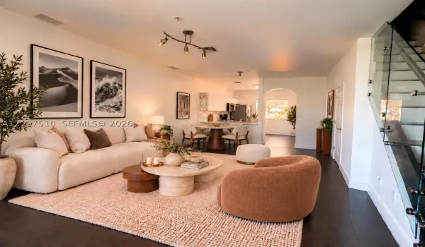 Open-concept interior featuring dark-tone wood-finish flooring, a contemporary multi-bulb ceiling fixture, a glass-panel staircase railing, and a kitchen area with white cabinetry and an arched entryway