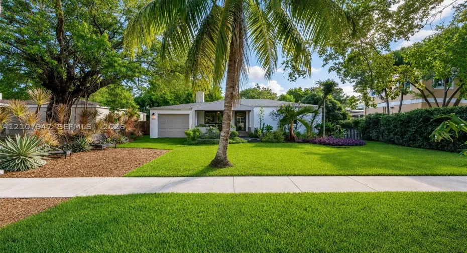 Single-story residence featuring a light-toned exterior, an attached one-car garage, manicured lawn, palm tree, and mature landscaping