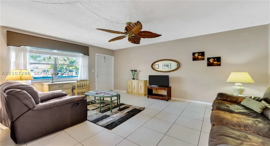 Neutral-toned interior featuring oversized tile flooring, a large window with a built-in cornice, and a ceiling fan with decorative leaf-shaped blades
