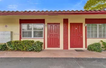Exterior facade featuring red panel doors, grid-pattern windows, terra-cotta style roofing tiles, and a concrete pathway with low-profile shrubbery