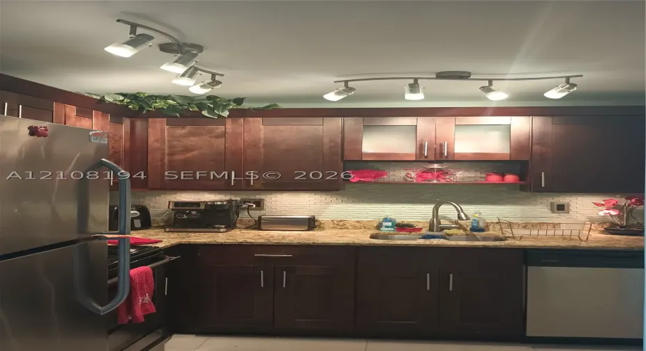 Dark wood cabinetry featuring brushed metal hardware, stone-finish countertops, textured tile backsplash, stainless steel appliances, and dual adjustable track lighting fixtures