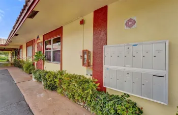 Covered exterior walkway featuring stucco walls, a multi-unit metal mailbox system, and established shrubbery