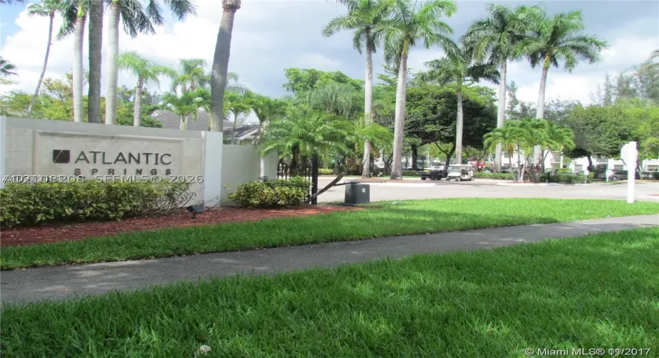 Community entrance featuring stone-clad monument signage, manicured turf, mature palms, and paved roadways